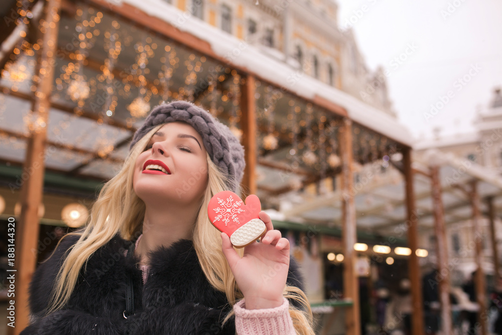 Fototapeta premium Positive blonde model with closed eyes holding palatable gingerbread cookie at the light decoration at the square in Kyiv