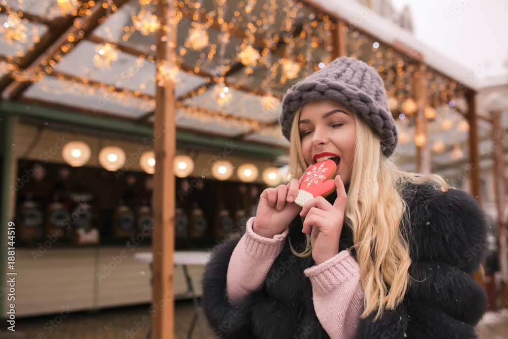 Amazing blonde girl bites tasty gingerbread cookie against light decoration at the square in Kiev
