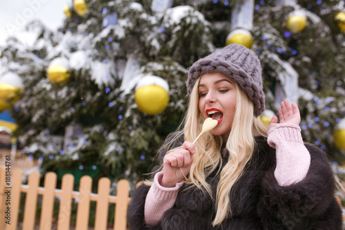 Beautiful blonde woman in grey knitted hat bites delicious chocolate candy against Christmas spruce