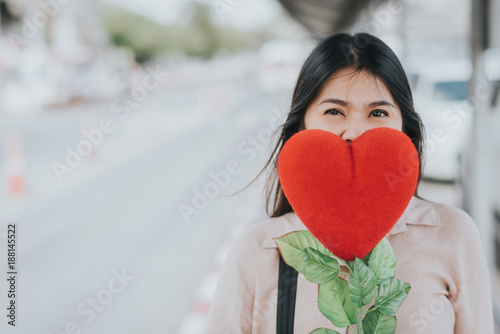 Attractive young Asian woman holding red love hearts flower over her mouth outdoor