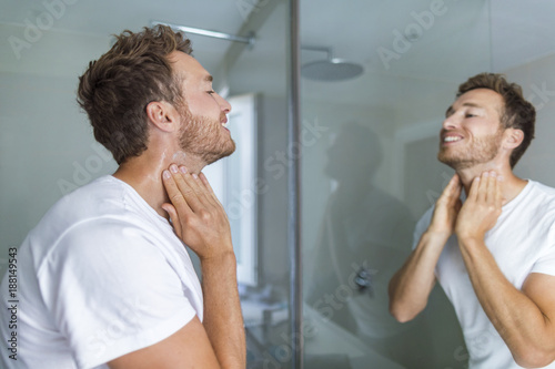 Man putting after shave perfume lotion or skin care cream for sensitive skin after shaving beard looking in bathroom mirror on neck. Male beauty, beard care skincare concept. photo