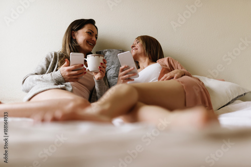 Gorgeous friends using the mobile and chatting in the bedroom.