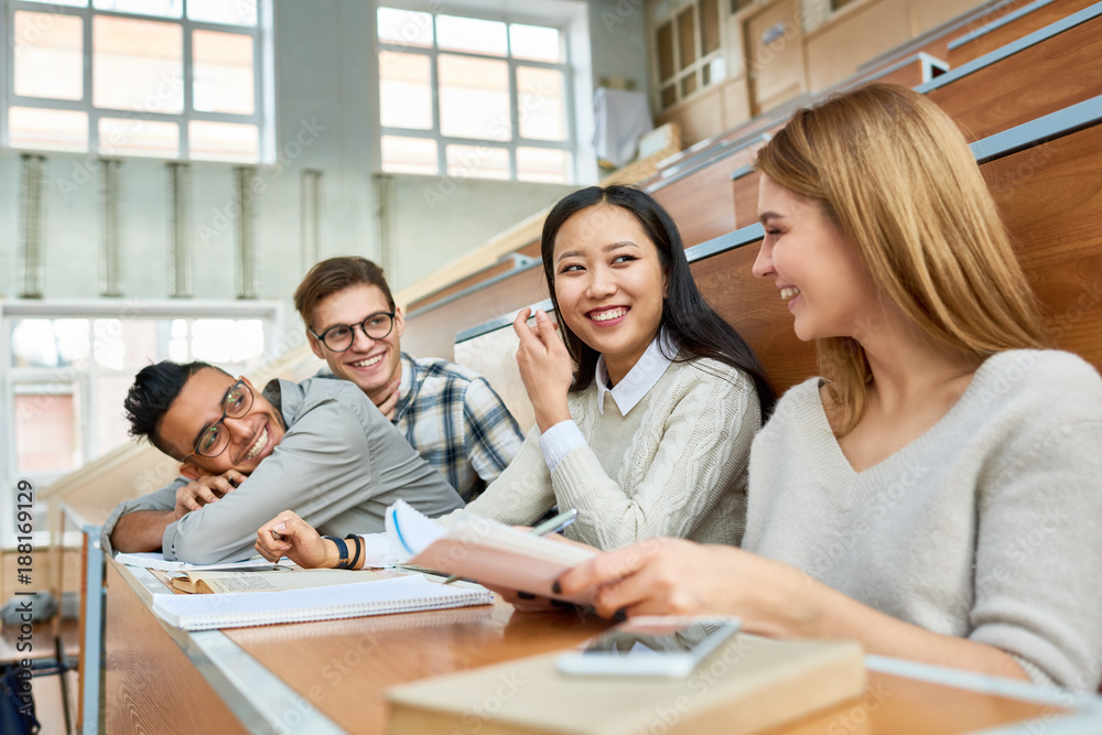 Multi-ethnic group of cheerful students sitting at desks in lecture hall of modern college and laughing, focus on two beautiful girls in front Multi-ethnic group of cheerful students sitting at desks in lecture hall of modern college and laughing, focus on two beautiful girls in front