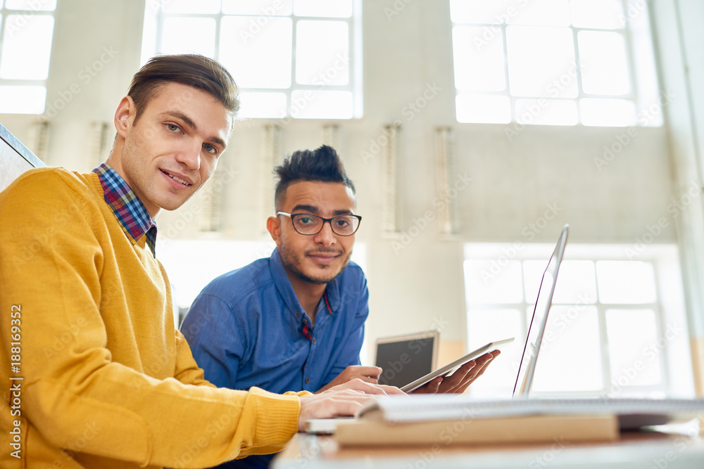 Portrait of two smiling students sitting at desk in modern lecture hall at college and looking at camera, one of them Middle-Eastern Portrait of two smiling students sitting at desk in modern lecture hall at college and looking at camera, one of them Middle-Eastern