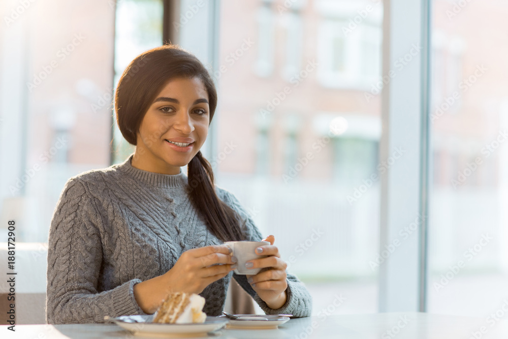 Obraz premium Young woman with cup of coffee and cake looking at camera while sitting in cafe