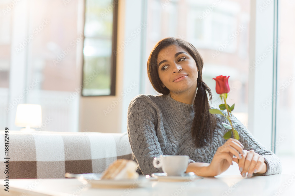 Happy young woman with fresh rose sitting by table in cafe and looking aside while thinking about her sweetheart Happy young woman with fresh rose sitting by table in cafe and looking aside while thinking about her sweetheart