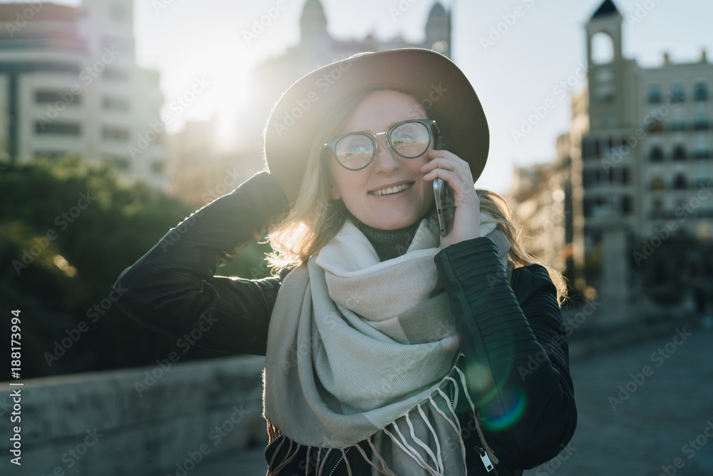 Sunny autumn day, backlight. Young attractive woman tourist in hat and eyeglasses stands on city street, talking on cell phone, smiling, laughing. Hipster girl walks. Vacation, adventure, trip.