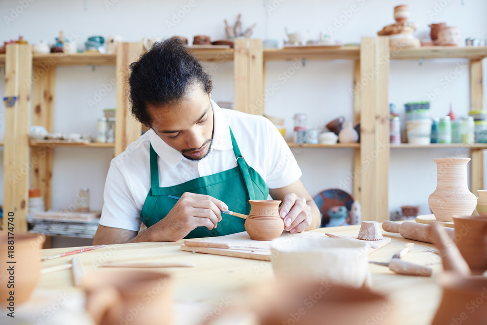 Young creative man with paintbrush painting earthenware by workplace