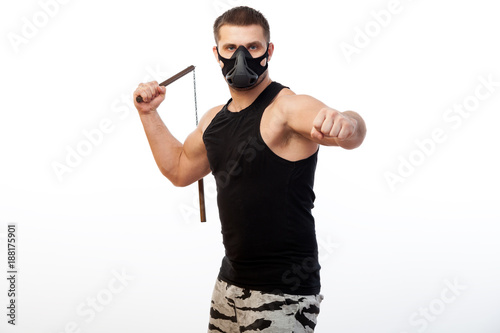 A young sporty blue-eyed man in a sporty black T-shirt, wearing boxing gloves and a black training mask is holding a metal nunchak in his hand on a white isolated background photo