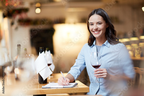 Happy young sommelier with glass of red wine tasting it and making notes photo