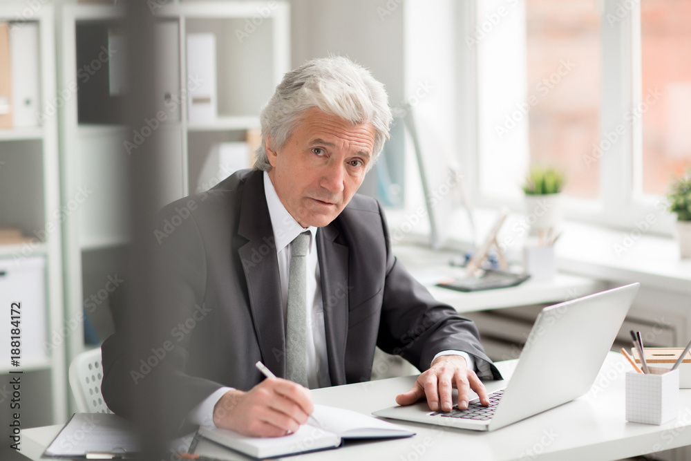 Experienced director in formalwear sitting by workplace, networking and making notes
