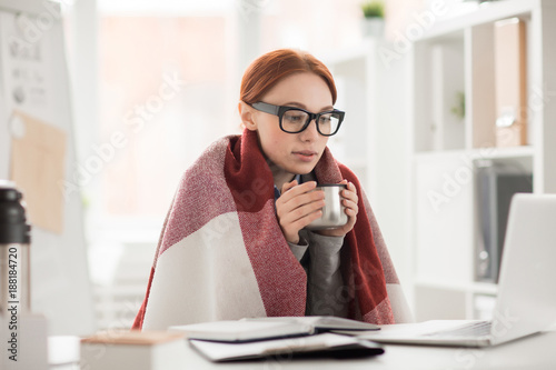 Young woman in plaid and eyeglasses drinking hot tea from mug during work in office
