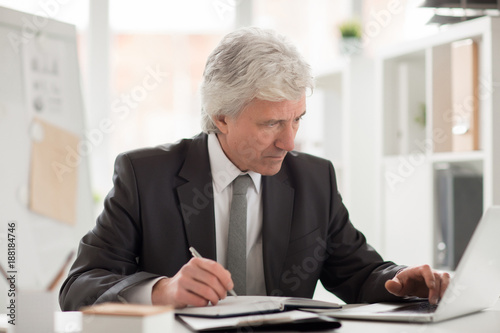 Serious boss in suit sitting by workplace with laptop in front and reading online data