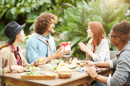 Young man giving box with present to his girlfriend by festive table with their friends near by