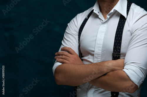 Hands of an unidentified strong man in formal attire with suspenders folded on chest on blue grunge background