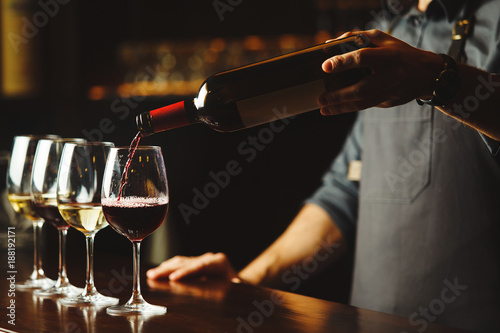 Fototapeta Bartender pours red wine in glasses on wooden bar counter