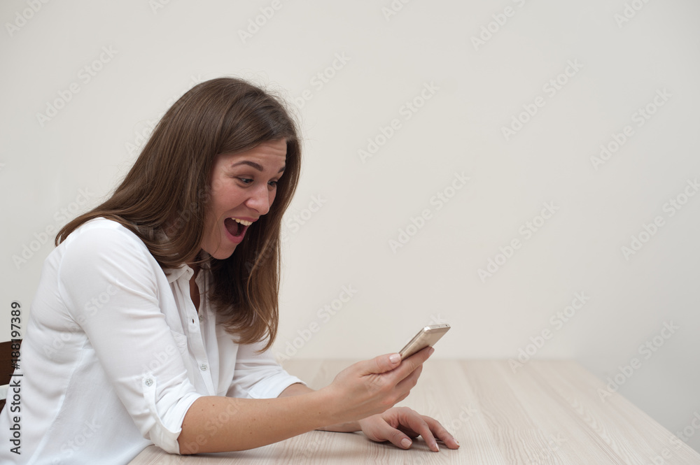 Portrait of beautiful shocked young woman sitting at the table with smartphone and looking at the screen on white wall background. Copyspace Portrait of beautiful shocked young woman sitting at the table with smartphone and looking at the screen on white wall background. Copyspace