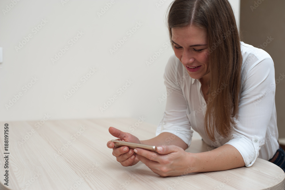 Portrait of beautiful young woman using her smartphone sitting at the table on white wall background Portrait of beautiful young woman using her smartphone sitting at the table on white wall background