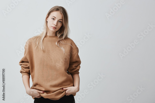 Cute pretty beautiful woman wears loose sweater and black trousers, holds her hands in pocket, looks with appeal at camera. Pleasant-looking girl posing against gray background photo