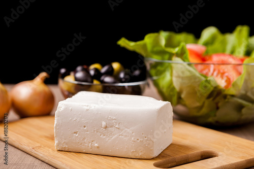 Close up on Feta cheese on wooden board on a table with summer salad, onions and glass bowl filled with olives