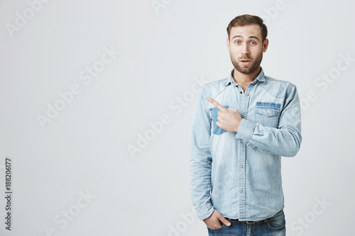 Amazed shocked male model with beard, wearing denim shirt, looking in surprisement at camera with opened mouth, pointing with forefinger at copy space for your text or advertisement