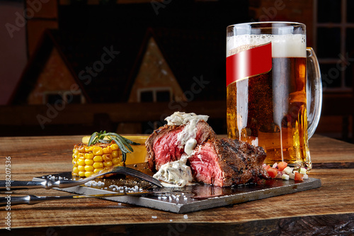 Grilled steak with corn with mushrooms sauce on cutting board and mug of beer on wooden table
