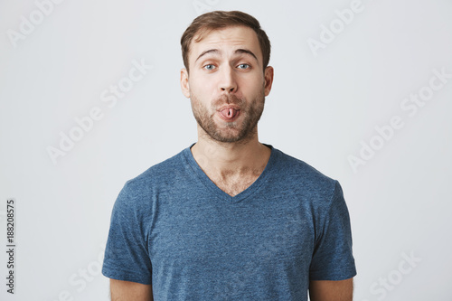 Gloomy young male with beard showing his digust and distaste having gloomy expression demonstrating tongue refusing to do something. Making faces handsome man with rejecting guilt isolated on gray