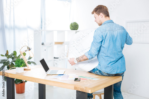 Important graphic. Clever serious responsible engineer looking attentively at the big drawing in his hands and sitting on the table while thinking about his project