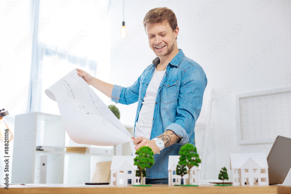 Beautiful project. Cheerful smart skilled engineer looking satisfied while standing near the table and holding a big detailed drawing in his hands Beautiful project. Cheerful smart skilled engineer looking satisfied while standing near the table and holding a big detailed drawing in his hands