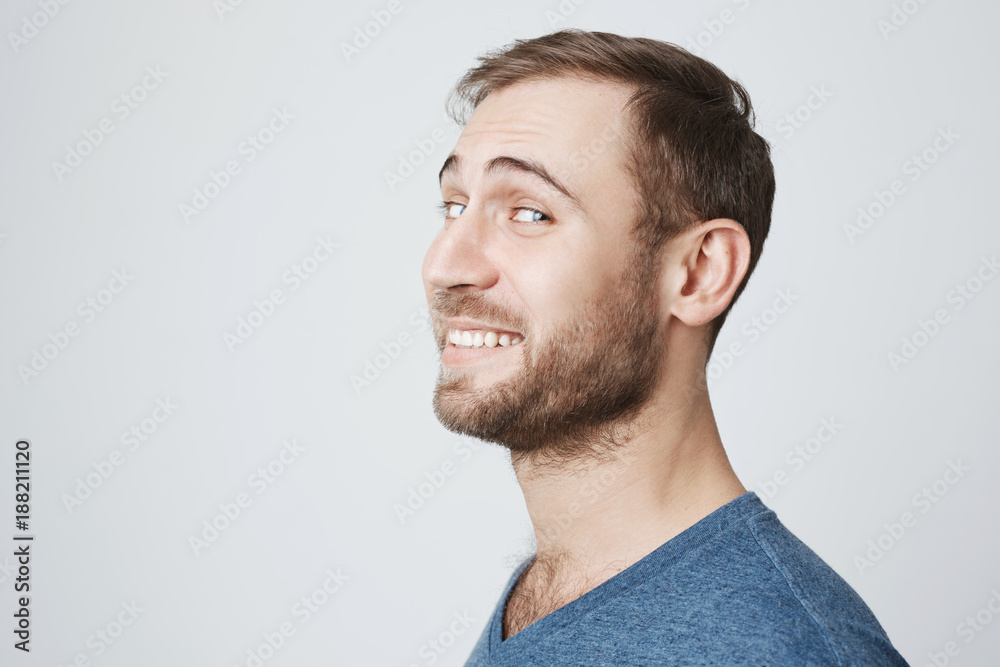Obraz premium Profile of smiling good-looking attractive man in blue t-shirt with stubble and dark hair, looking at camera, expressing positive emotions, posing against gray studio wall.