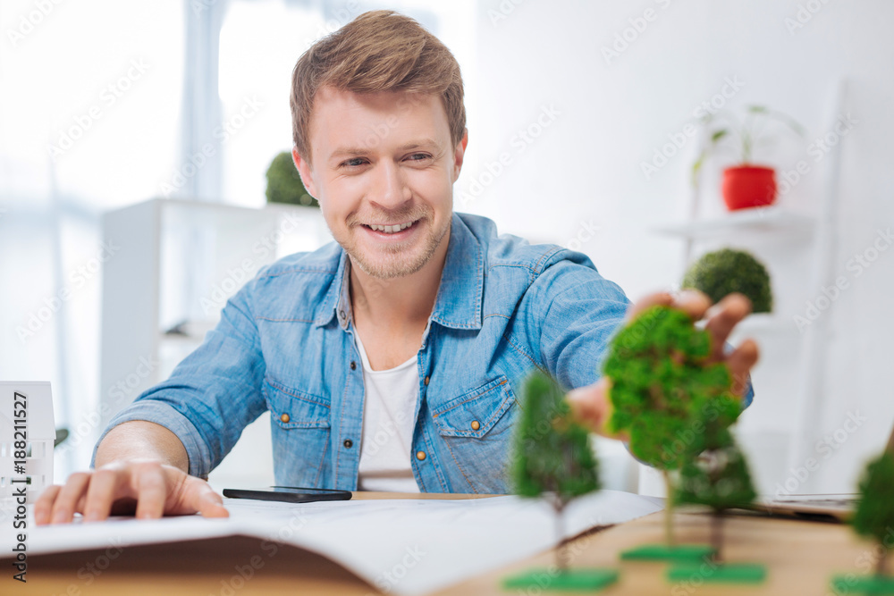 Amazing material. Positive emotional young engineer looking impressed by the modern materials while sitting at the table in his comfortable office and touching a miniature tree Amazing material. Positive emotional young engineer looking impressed by the modern materials while sitting at the table in his comfortable office and touching a miniature tree