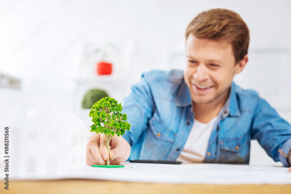 Pretty tree. Cheerful emotional young engineer feeling excited while being in his office and looking at the adorable realistic miniature tree and touching it Pretty tree. Cheerful emotional young engineer feeling excited while being in his office and looking at the adorable realistic miniature tree and touching it