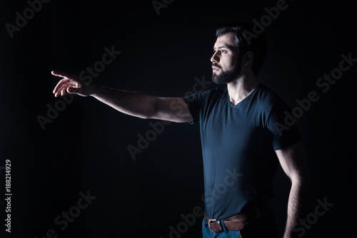 Brutal look. Handsome nice serious man looking in front of him and pointing with his hand while standing against black background