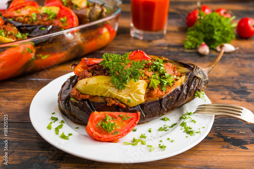 Eggplants stuffed with minced meat and cooked with fresh tomatoes and bell peppers on white plate and in baking dish on wooden rustic table.