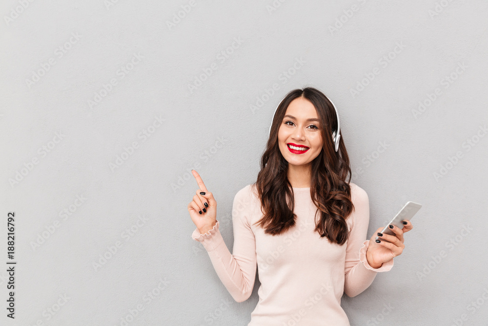 Horizontal photo of beautiful female in wireless headphones holding mobile phone pointing index finger aside over gray background copy space