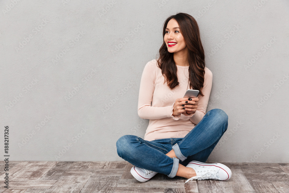 Portrait of smiling woman with red lips typing text message or scrolling in internet using mobile phone sitting in lotus pose over gray background Portrait of smiling woman with red lips typing text message or scrolling in internet using mobile phone sitting in lotus pose over gray background