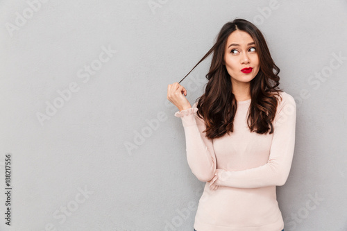 Portrait of young tricky woman touching lock of hair meaning she has something in mind with facial expressions over gray background