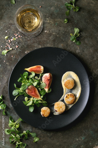 Fried scallops with lemon, figs, sauce and green salad served on black plate with glass of white wine over old dark metal background. Top view, space. Plating, fine dining