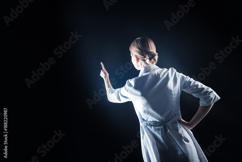 Futuristic technology. Professional nice female scientist standing against black background and holding her hand up while using transparent screen