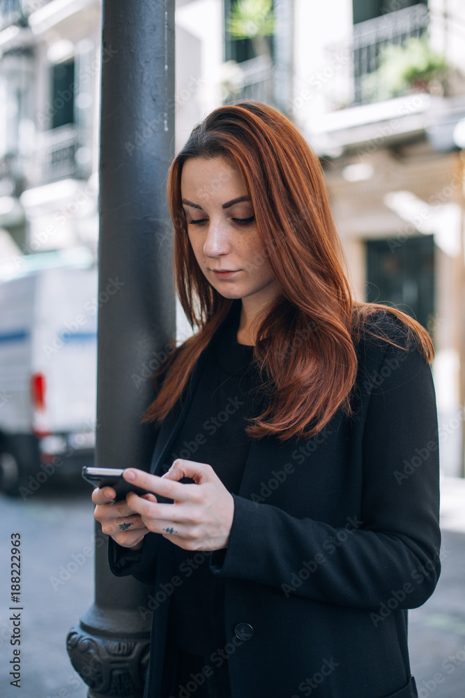 Beautiful casual woman with red hair and natural makeup stands on street and texts or chats on her smartphone, connects with friends and family, vertical mobile screen verison Beautiful casual woman with red hair and natural makeup stands on street and texts or chats on her smartphone, connects with friends and family, vertical mobile screen verison