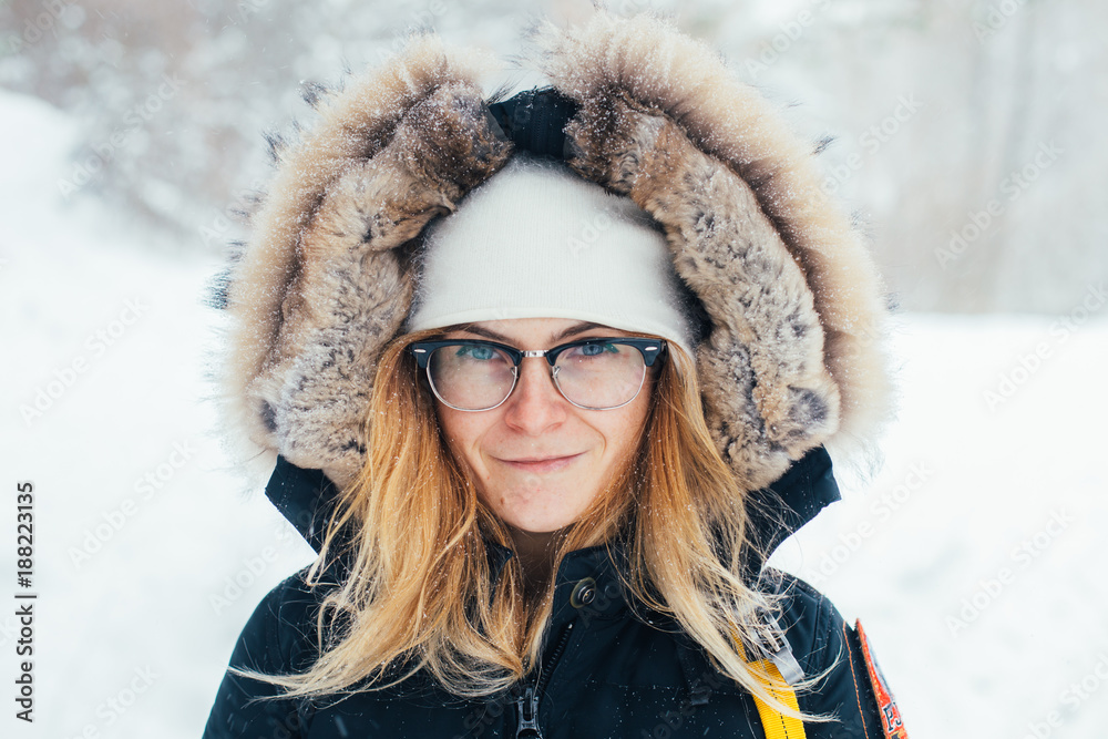 Close up portrait of beautiful smiling young woman with blond hair and glasses, wears huge jacket hood with fur and white hat. Looks at camera, in middle of snowy winter forest on cold day Close up portrait of beautiful smiling young woman with blond hair and glasses, wears huge jacket hood with fur and white hat. Looks at camera, in middle of snowy winter forest on cold day
