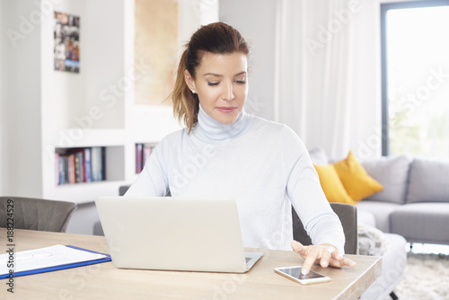 Free Download Managing my business from home. A beautiful middle aged woman using laptop and mobile phone while sitting at desk and working from home. Home office.  - GFXLoad