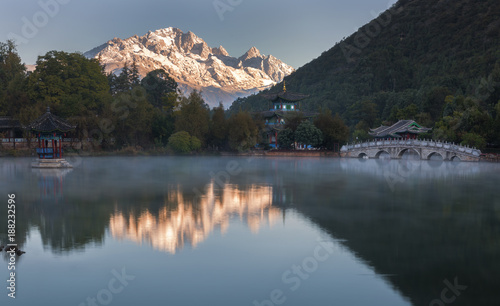 Chinese pagoda and moon bridge surround in forest with reflection in foggy lake with first sun ray touch snow mountain range background in morning at Black Dragon pool, Lijiang, China