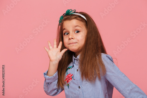 Portrait of funny female kid having long auburn hair looking on camera giving high five meaning hi or bye with hand over pink background