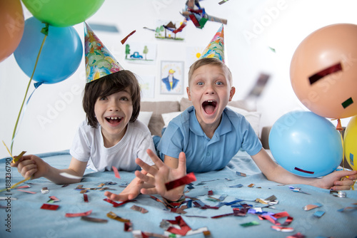 Portrait of two jolly youngsters in living room holding colorful balloons, children are surrounded with confetti