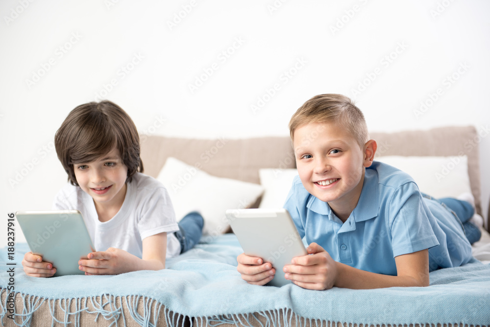These gadgets are awesome. Portrait of enjoyed classmates spending holidays at home. They are holding devices while resting on sheets These gadgets are awesome. Portrait of enjoyed classmates spending holidays at home. They are holding devices while resting on sheets