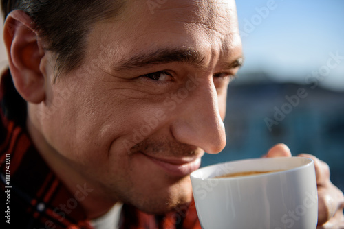 Close up portrait of contented guy looking at camera with smile, he is having a cup of drink in his hands. Focus on his face