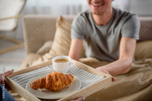 Close up of male and female arms holding tray, focus on salver with coffee and croissant. Man is smiling and sitting in bed
