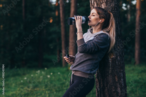 Female jogger recovering after intensive workout standing near the tree drinking water, wearing headphones in park