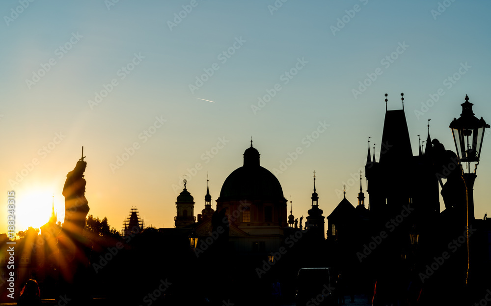 From Charles Bridge before sunrise looking into rising sun silhouettes of rooftops, domes and skyline backlit by golden glow.